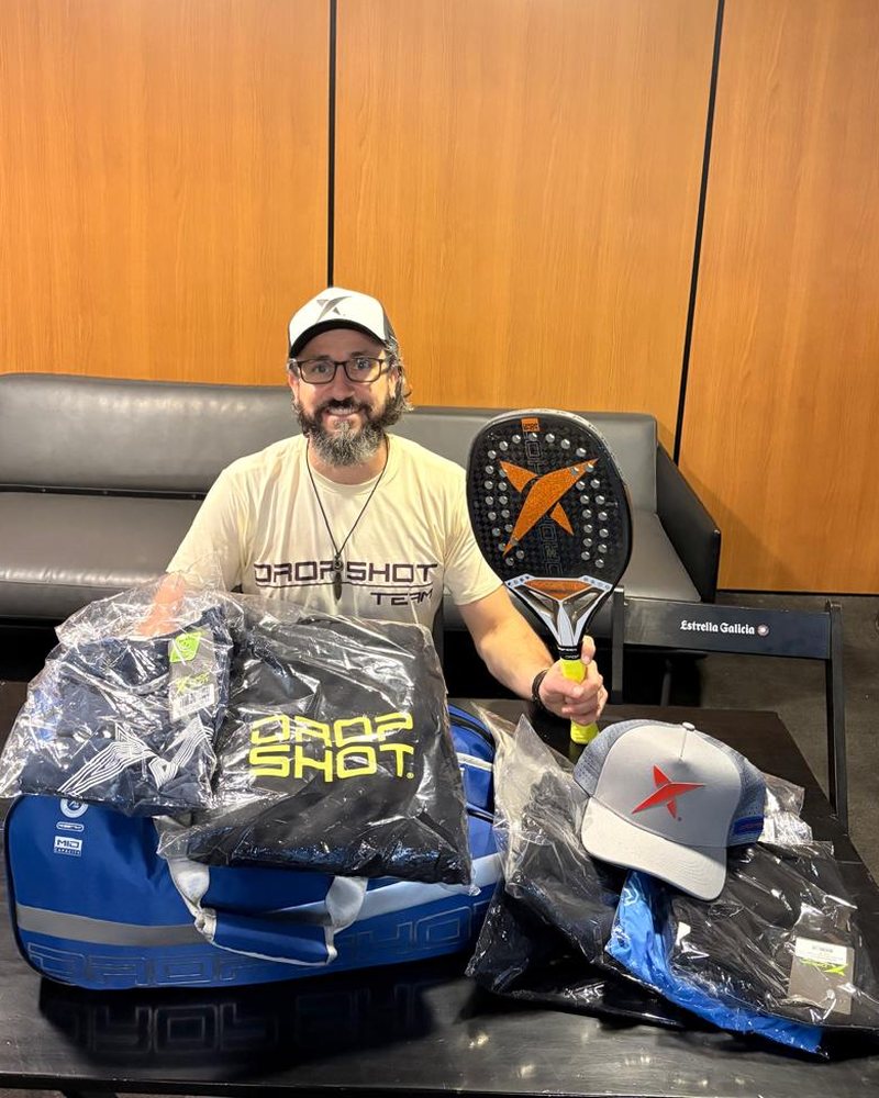 Formiga sorrindo, de boné e camiseta Drop Shot Team, segurando uma raquete Drop Shot, com produtos da marca sobre a mesa